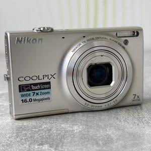 Può includere: Una fotocamera digitale Nikon Coolpix color argento con obiettivo Nikkor 7x wide optical zoom ED VR con escursione focale 5.0-35.0mm 1:3.7-5.6. Il corpo anteriore riporta le scritte 16.0 Megapixels, Touch Screen e 7x Wide, con flash integrato.