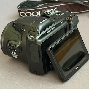 May include: A black Nikon Coolpix L830 digital camera with a tilting rear LCD screen. The camera body features a 34x optical zoom lens, a top-mounted power button, and a dedicated video recording button. A black camera strap with white text is included.