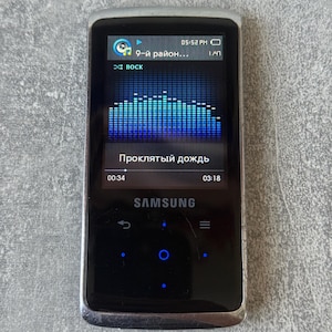 May include: A silver and black Samsung digital media player with a rectangular screen displaying a blue audio equalizer, the text 9-й район, the word ROCK, and the song title Проклятый дождь, featuring touch-sensitive navigation buttons below.
