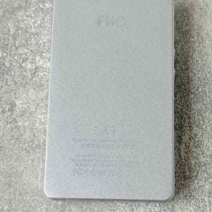 May include: A rectangular silver FiiO X1 portable high-resolution music player with a metallic finish. The device features the FiiO logo at the top, the X1 model name in the centre, and technical text printed at the bottom including FCC markings.