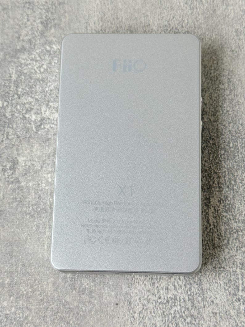 May include: A rectangular silver FiiO X1 portable high-resolution music player with a metallic finish. The device features the FiiO logo at the top, the X1 model name in the centre, and technical text printed at the bottom including FCC markings.