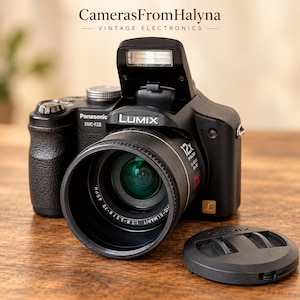 Peut inclure: Un appareil photo numérique noir Panasonic Lumix DMC-FZ8 avec un flash escamotable, un zoom optique 12x et un bouchon d'objectif. Le boîtier présente le logo Lumix, le numéro de modèle et les spécifications de l'objectif. Le texte CamerasFromHalyna Vintage Electronics est visible au-dessus.