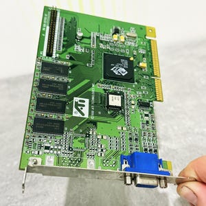 Op de afbeelding: Een groene ATI Technologies grafische kaart met een opvallende zwarte 3D Rage LT PRO chip, meerdere geheugenmodules en een blauwe VGA-poort gemonteerd op een zilveren metalen beugel. De printplaat toont diverse elektronische componenten en tekst.