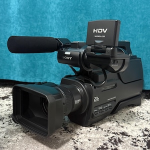 Puede incluir: Videocámara Sony HDV negra con pantalla LCD ancha y lente de zoom 20x. Cuenta con un micrófono con cubierta de espuma negra y la inscripción "Power SteadyShot". La videocámara está sobre una superficie estampada.