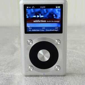 May include: A silver digital music player with a rectangular colour screen displaying the text addiction doubt the dosage and 03. Addiction Crew - Download.mp3. The device features a large circular black control wheel with a central silver button.