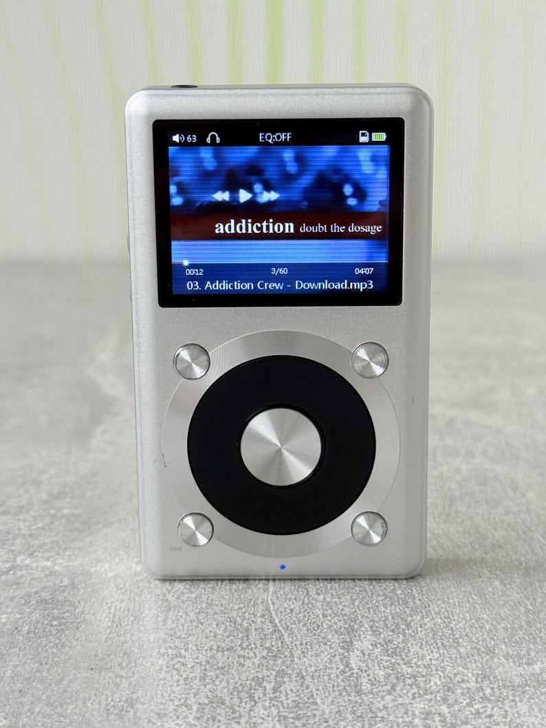 May include: A silver digital music player with a rectangular colour screen displaying the text addiction doubt the dosage and 03. Addiction Crew - Download.mp3. The device features a large circular black control wheel with a central silver button.