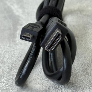Puede incluir: Un cable HDMI negro con un conector micro-HDMI y un conector HDMI estándar. El cable está enrollado y asegurado con una tira de velcro negra. El texto "WITH ETHERNET" está impreso en blanco sobre la cubierta negra del cable.