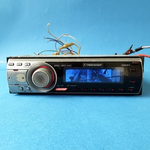 May include: A Pioneer car stereo with a black face, silver trim, and a central control knob. The stereo has a display screen and various buttons. Wires extend from the back. The stereo is on a blue background.