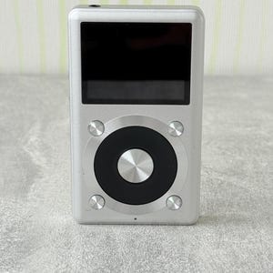 May include: A silver digital music player with a rectangular black screen and a circular control interface. The device features a central metallic button surrounded by a black ring, with four smaller silver buttons positioned at the corners.