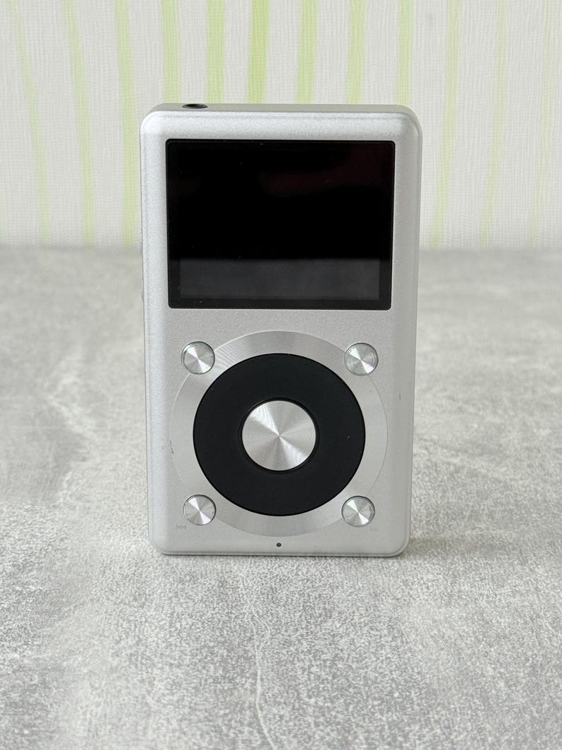 May include: A silver digital music player with a rectangular black screen and a circular control interface. The device features a central metallic button surrounded by a black ring, with four smaller silver buttons positioned at the corners.