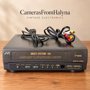 Pode incluir: Um leitor de cassetes de vídeo VHS multissistema JVC preto com ranhura de carregamento frontal, botões de controlo e visor de seguimento automático digital. A unidade apresenta texto como Multi System, P28 e Made in Japan com cabos.