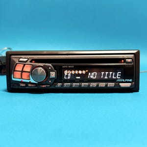 May include: Black car stereo with orange and black buttons and a digital display showing "NO TITLE". The stereo has the brand name "ALPINE" and supports MP3/WMA formats. The model number is COE-9827RM.