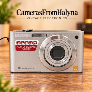 Puede incluir: Una cámara digital Lumix plateada con una etiqueta de 10 mega pixels, un objetivo Lumix DC Vario con zoom óptico de 4x y una pegatina roja que muestra iconos de Auto Scene Mode. El cuerpo tiene un acabado metálico y un pequeño logotipo L dorado.