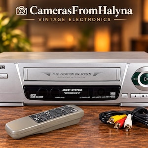 Könnte beinhalten: Ein silberner JVC VHS-Videokassettenrekorder mit einem Frontlader-Kassettenschacht mit der Aufschrift TAPE POSITION ON-SCREEN, einer runden grünen Wiedergabetaste und einem schwarzen Display. Enthält eine passende graue Fernbedienung sowie gelbe, rote und weiße Cinch-Kabel.