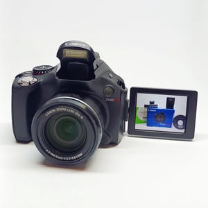 May include: A black Canon SX40 HS digital camera with a built-in flash and a 4.3-150 mm lens. The camera has a swiveling LCD screen that is displaying a picture of a Canon camera.