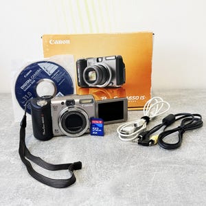 May include: A silver and black Canon PowerShot A650 IS digital camera with a flip-out screen, a 512MB SD memory card, a white USB cable, a black AV cable, a software disc, and the original orange and white product box with camera imagery.