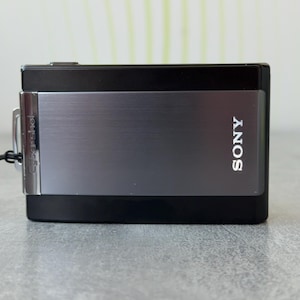 Op de afbeelding: Een rechthoekige Sony Cyber-shot digitale camera met een strakke zwarte behuizing en een geborsteld metallic grijs middenpaneel. De merknaam Sony staat in het wit aan de rechterkant, terwijl het Cyber-shot logo aan de linkerkant zichtbaar is.