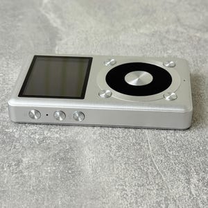 May include: A silver digital music player featuring a rectangular screen on the left and a circular control interface on the right. The device has three circular buttons on the side edge, including a power button and volume controls.