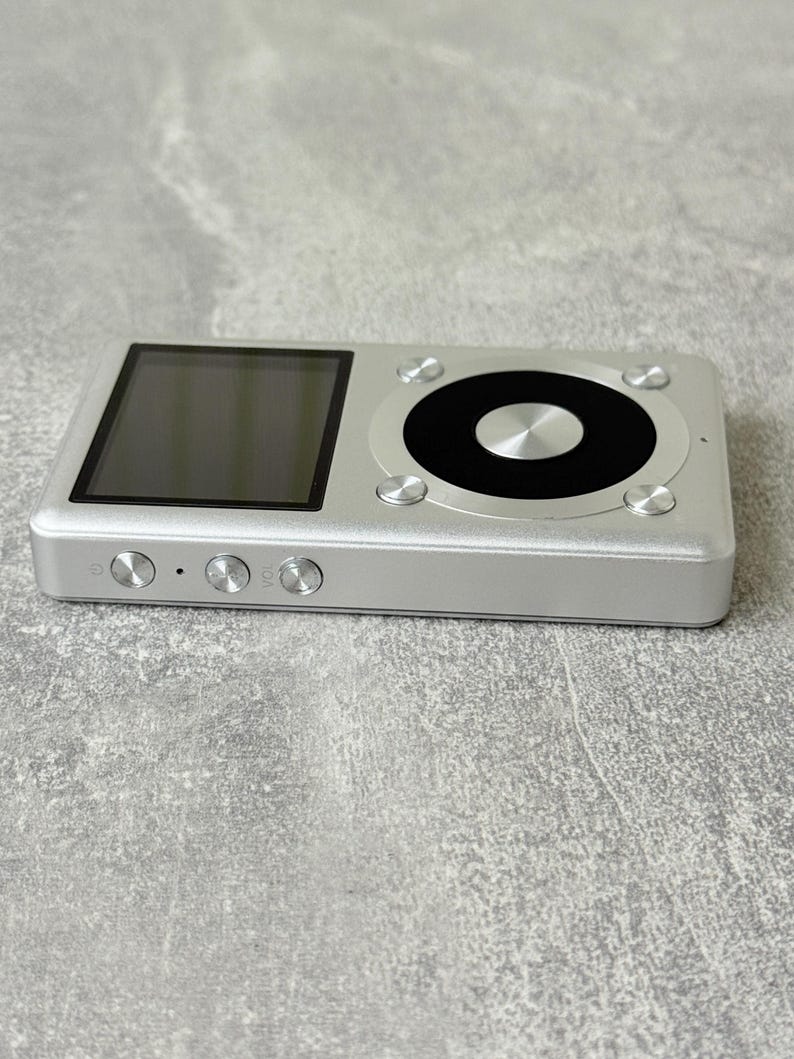 May include: A silver digital music player featuring a rectangular screen on the left and a circular control interface on the right. The device has three circular buttons on the side edge, including a power button and volume controls.