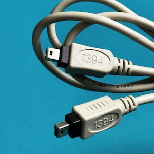 Może przedstawiać: Zbliżenie dwóch kabli FireWire 1394 z szarymi złączami i jasnoszarym kablem na niebieskim tle. Na złączach wydrukowano numer "1394".