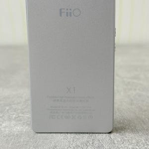 May include: A silver rectangular FiiO X1 portable high-resolution music player. The device features the FiiO logo at the top, the model name X1 in the centre, and technical specifications and regulatory icons printed at the bottom.