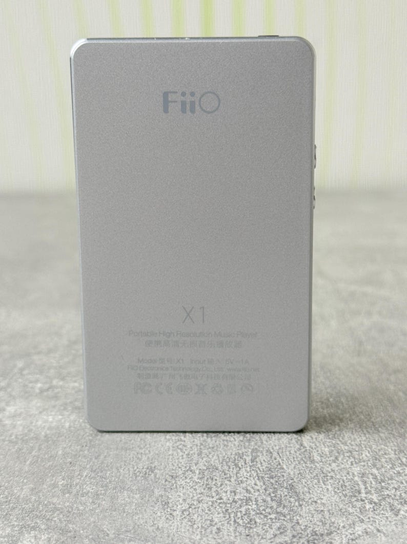 May include: A silver rectangular FiiO X1 portable high-resolution music player. The device features the FiiO logo at the top, the model name X1 in the centre, and technical specifications and regulatory icons printed at the bottom.