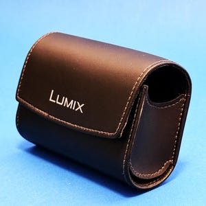 Puede incluir: Una funda de cámara de cuero negro con la palabra "LUMIX" impresa en blanco. La funda tiene un cierre de solapa y un borde cosido. La funda está sobre un fondo azul.