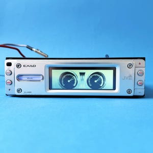Puede incluir: Autorradio plateado con una pantalla digital que muestra dos medidores circulares. El panel frontal tiene botones etiquetados como "OK" y "VOL/AC". Las marcas "EXAD" y "JVC" son visibles. El número de modelo KD-LX501 está impreso en la cara.