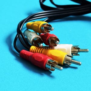May include: A selection of RCA audio/video cables with red, yellow, white, and grey connectors. The cables are black and set against a bright blue backdrop. The connectors are made of metal.
