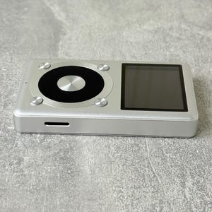 May include: A rectangular silver digital music player featuring a black square display screen on the right and a circular black control wheel on the left. The device has a metallic finish with four small silver circular buttons surrounding the wheel.