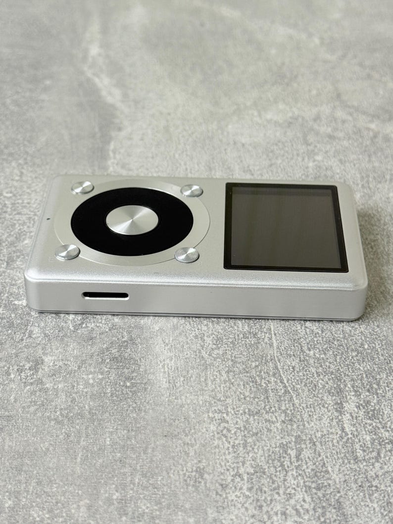 May include: A rectangular silver digital music player featuring a black square display screen on the right and a circular black control wheel on the left. The device has a metallic finish with four small silver circular buttons surrounding the wheel.