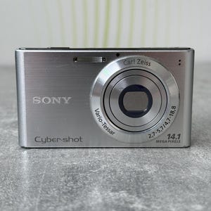 May include: A silver Sony Cyber-shot digital camera featuring a Carl Zeiss Vario-Tessar lens with 2.7-5.7/4.7-18.8 specifications and 14.1 mega pixels text printed on the front face. The compact device has a metallic finish and a rectangular body.