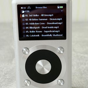 May include: A silver digital music player with a rectangular screen displaying a file list including 3rd Strike, 40 Below Summer, Addiction Crew, Blindspott, Boiler Room, and Catatonik. It features a circular black control wheel with a silver centre.