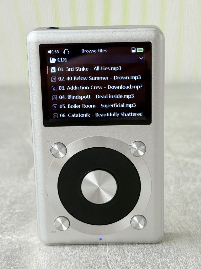 May include: A silver digital music player with a rectangular screen displaying a file list including 3rd Strike, 40 Below Summer, Addiction Crew, Blindspott, Boiler Room, and Catatonik. It features a circular black control wheel with a silver centre.