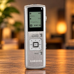 May include: A silver Samsung digital voice recorder with a vertical rectangular body, a monochrome LCD screen displaying "VOICE", "008", "Stand by", and "0:02:18", a central navigation pad with playback controls, and a brushed metal finish.