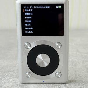 May include: A silver digital music player with a rectangular screen displaying a language selection menu. The device features a large circular black control wheel with a central silver button and four smaller silver buttons at the corners.