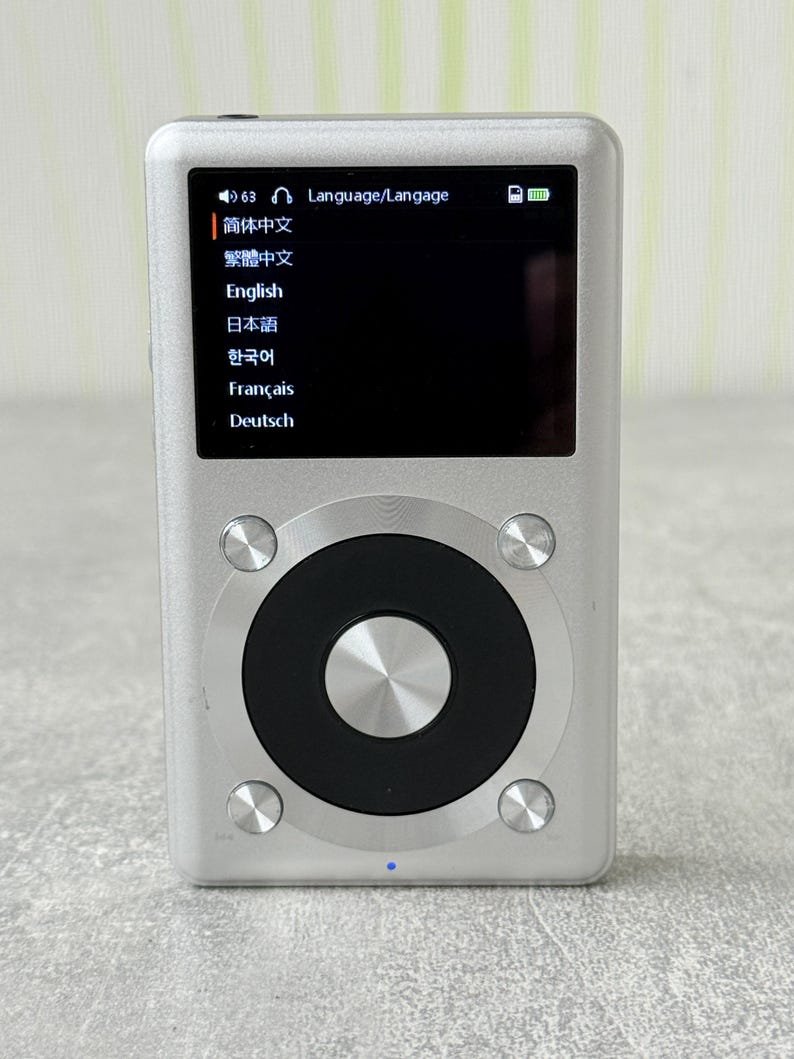 May include: A silver digital music player with a rectangular screen displaying a language selection menu. The device features a large circular black control wheel with a central silver button and four smaller silver buttons at the corners.