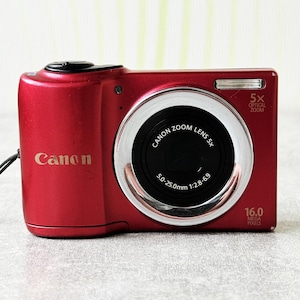 Puede incluir: Cámara digital Canon roja con objetivo zoom óptico 5x. La cámara tiene una lente negra y un anillo plateado alrededor de la lente. La cámara tiene el texto "Canon" en la parte delantera y "16.0 MEGA PIXELS" en el lateral.