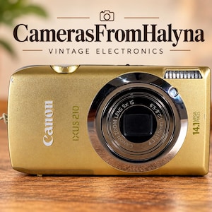 Peut inclure: Un appareil photo numérique Canon IXUS 210 doré doté d'un objectif zoom 5x et de 14,1 mégapixels. La face avant affiche le logo Canon, le nom du modèle IXUS 210 et les spécifications de l'objectif 4,3-21,5 mm 1:2,8-5,9. Une dragonne est fixée.