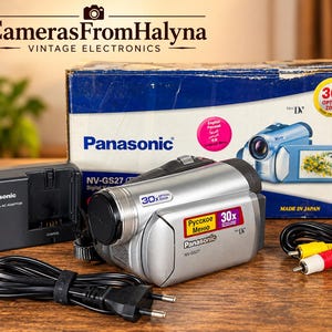 Puede incluir: Una videocámara digital Panasonic NV-GS27 plateada con lente de zoom óptico de 30x, acompañada de su caja de cartón azul y blanca original, un cargador de batería negro y un cable AV negro con conectores RCA amarillo y rojo para conexión.