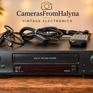 Peut inclure: Un lecteur VHS JVC noir avec les inscriptions B.E.S.T. Picture System, Auto Set et ShowView sur le panneau avant. L'appareil est accompagné d'une télécommande noire et de deux câbles de connexion noirs posés sur la surface supérieure.
