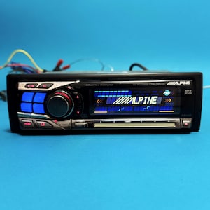 Puede incluir: Autorradio negro con botones y pantalla iluminados en azul. La radio tiene un mando central y la marca "ALPINE" es visible. Los cables se extienden desde la parte posterior de la radio.