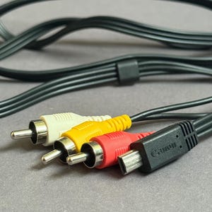 May include: A close-up of a black Canon AV cable with three RCA connectors in white, yellow, and red. The cable is black and has a black connector. The background is a light grey surface.