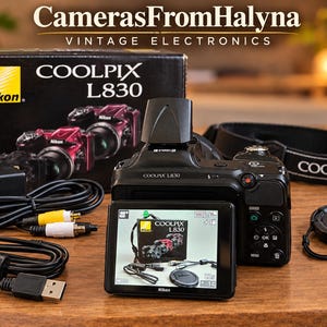 Op de afbeelding: Een zwarte Nikon Coolpix L830 digitale camera met een uitklapbaar scherm dat de camera zelf weergeeft. Inbegrepen zijn een zwarte doos met het Nikon-logo, een camerariem, een lensdop, een stroomadapter en twee zwarte kabels.