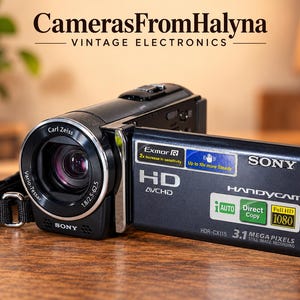 May include: A black Sony Handycam HDR-CX115 camcorder with a Carl Zeiss Vario-Tessar lens. The flip-out screen displays text including Exmor R, HD AVCHD, Sony, Handycam, iAUTO, Direct Copy, Full HD 1080, and 3.1 Mega Pixels still image recording.