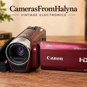 Puede incluir: Una videocámara HD Canon Legria HFR26 de color rojo oscuro con un objetivo de zoom avanzado de 28x. El dispositivo cuenta con una pantalla abatible que muestra el texto Canon, HD, 28x Advanced Zoom, Legria HFR26 y Built-in Memory 8 GB.