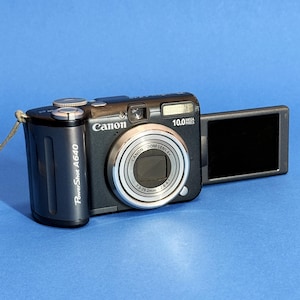 May include: A black Canon PowerShot A640 digital camera with a silver lens ring and a flip-out screen. The camera has a 10.0 Megapixels resolution and a 7.3-29.2mm zoom lens. The camera is on a blue background.
