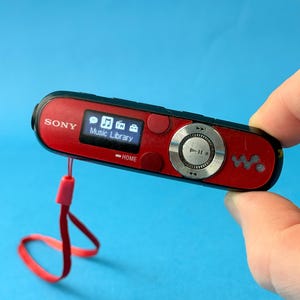 May include: A red and black Sony digital music player with a small screen displaying a music library interface. The device has a silver control wheel and a red lanyard. The word "SONY" is printed on the device.
