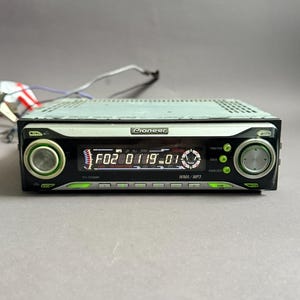 May include: A Pioneer car stereo with a black and silver faceplate. The stereo has a digital display showing numbers and text, with buttons and knobs on either side. Wires extend from the back.