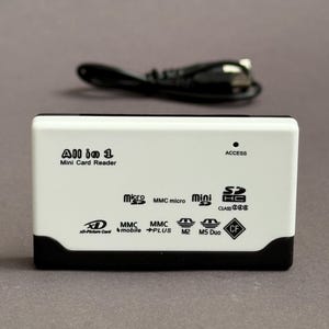 Peut inclure: Un lecteur de cartes "All in 1 Mini Card Reader" blanc et noir avec divers logos de types de cartes. L'appareil est accompagné d'un câble noir. Le texte "ACCESS" est visible.